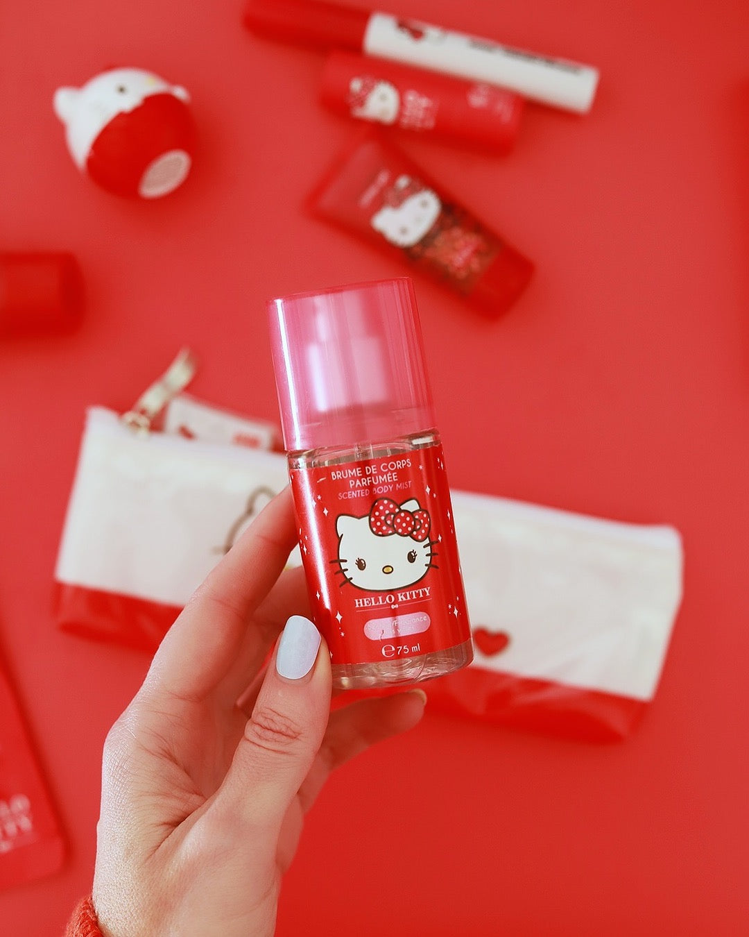 Une main aux ongles vernis de bleu clair présente le flacon de brume parfumée Hello Kitty au-dessus de trousses de beauté sur un fond rouge uni.