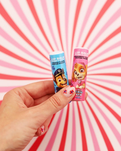 Une main tenant les deux tubes de baume à lèvres La Pat' Patrouille (Chase au parfum de biscuit et Stella au parfum de fraise). L'arrière-plan présente un motif dynamique de rayons rétro roses, blancs et rouges, et les ongles sont décorés de petites fleurs rouges.