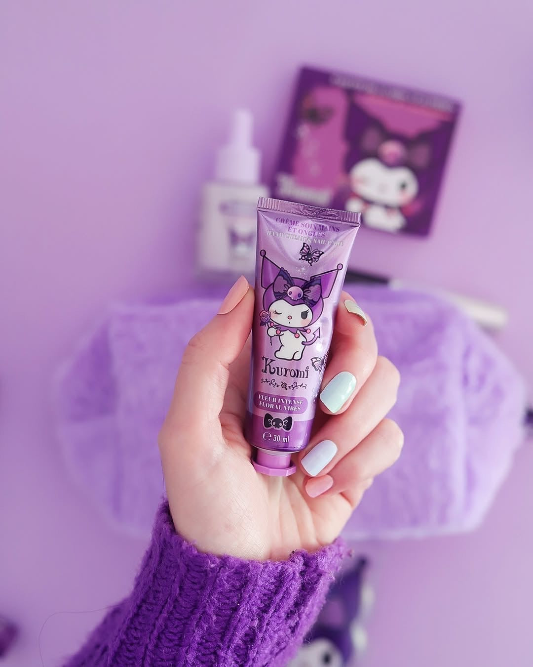 Modèle
Une main avec une manucure pastel tenant le tube de crème pour les mains et les ongles Kuromi Sanrio devant une trousse violette et les items de la collection Kuromi en arrière plan.