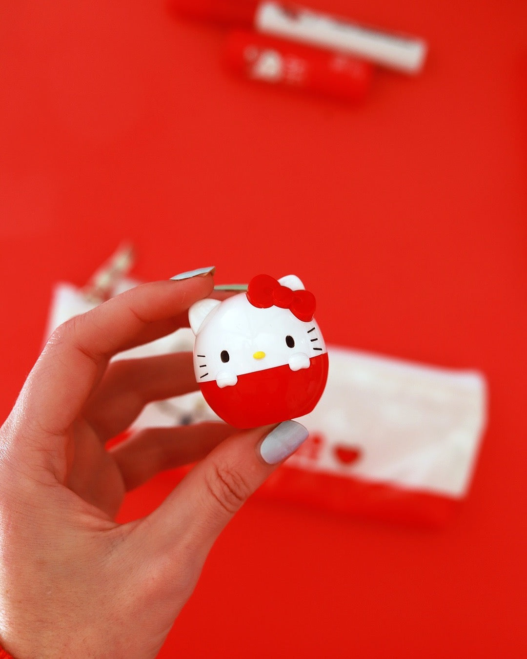 Une main tient le petit baume à lèvres œuf Hello Kitty rouge au-dessus d’une trousse de maquillage sur un fond rouge vif.