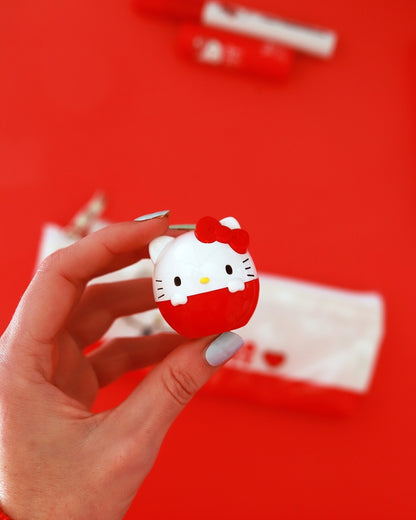 Une main tient le petit baume à lèvres œuf Hello Kitty rouge au-dessus d’une trousse de maquillage sur un fond rouge vif.