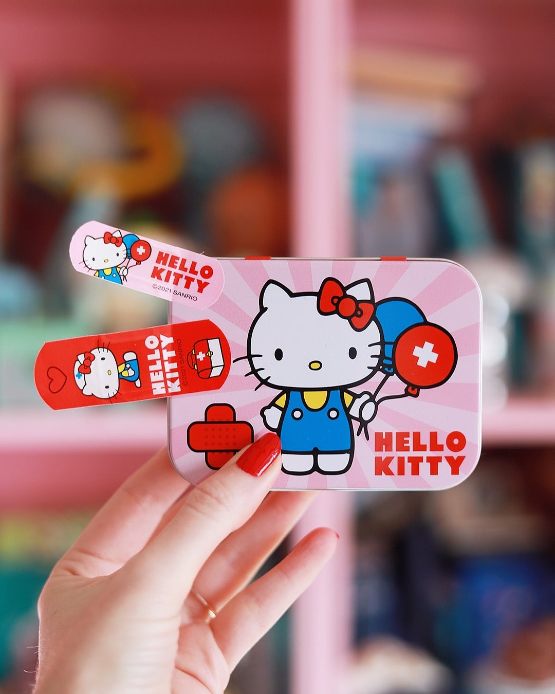 Une main aux ongles vernis en rouge tient la boîte métallique Hello Kitty ainsi que deux pansements individuels sur un fond rose flou.