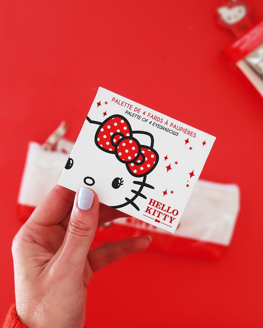Une main aux ongles vernis de bleu clair tient fermement le boîtier fermé de la palette Hello Kitty au-dessus d’une trousse sur un fond rouge vif.