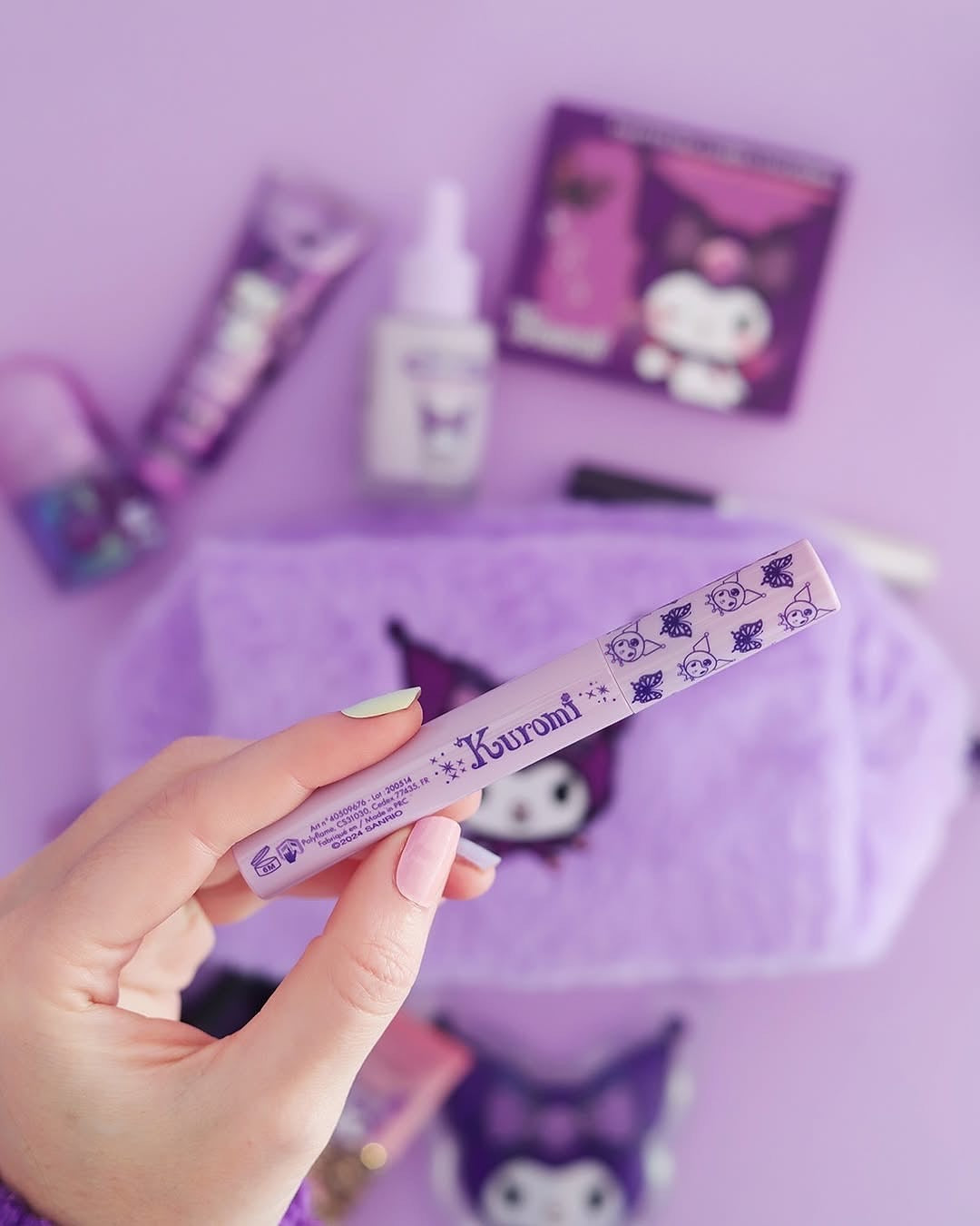 Une main avec une manucure violette et blanche tenant le tube de mascara noir volume Kuromi Sanrio devant une trousse en peluche et les produits de la collection en arrière plan.