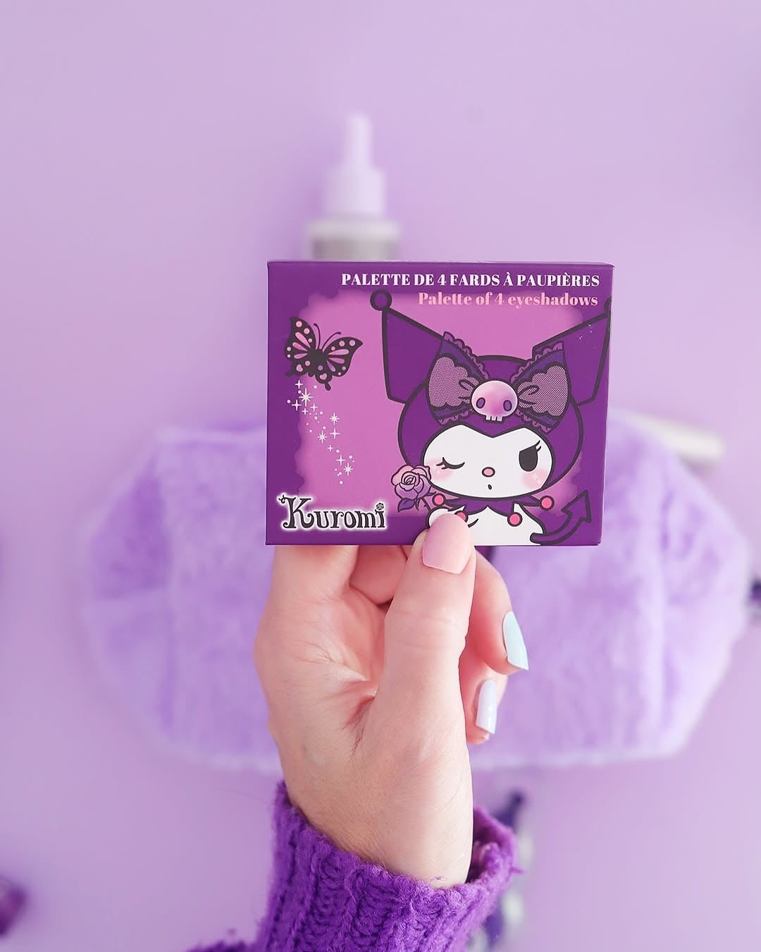 Une main présentant la boîte fermée de la palette de fards à paupières Kuromi Sanrio devant une ambiance d'un fond cosmétique violet.