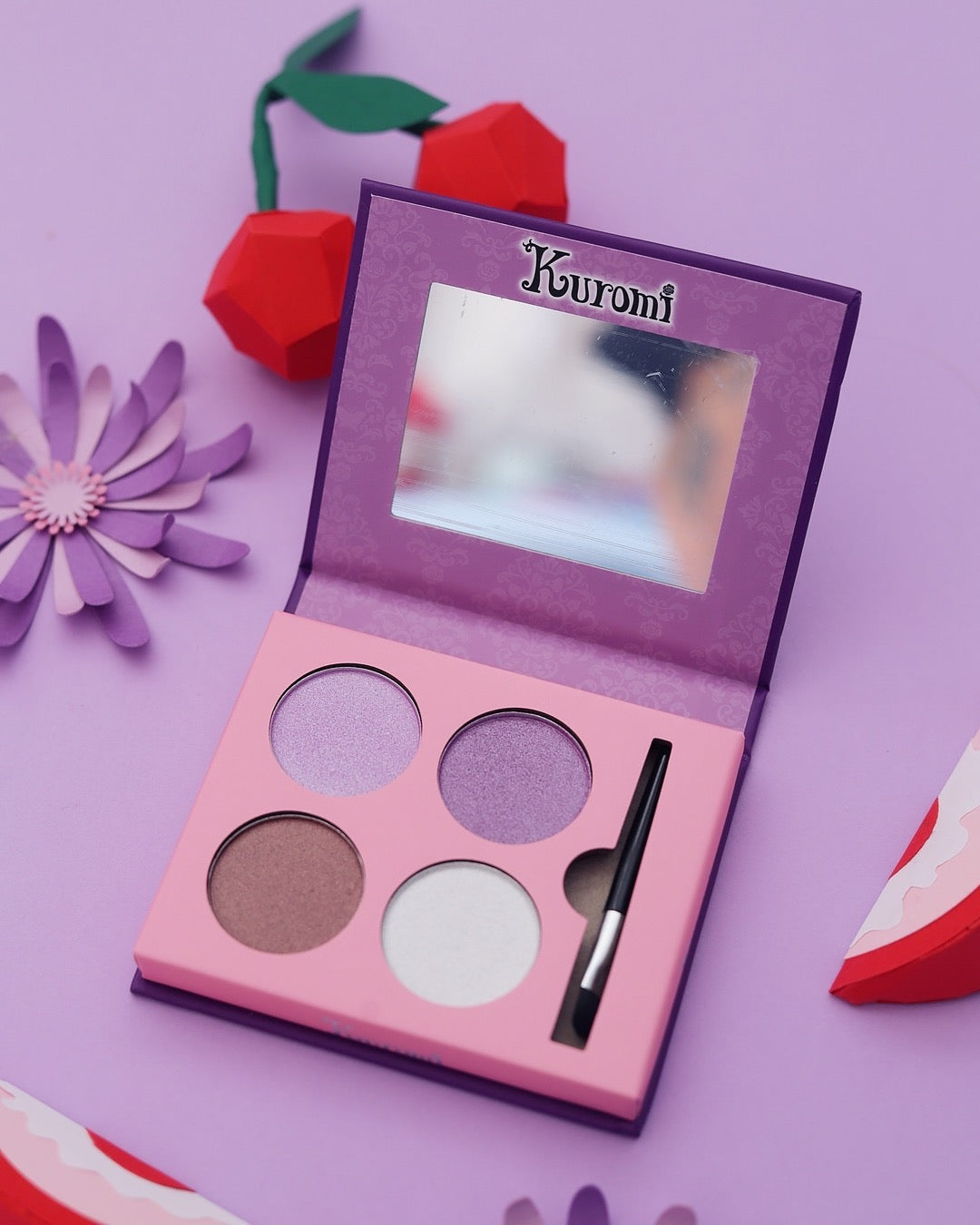 Palette de fards à paupières Kuromi ouverte montrant un miroir intégré, un pinceau applicateur et 4 teintes de fards (violet, mauve, brun et blanc). Dans une ambiance