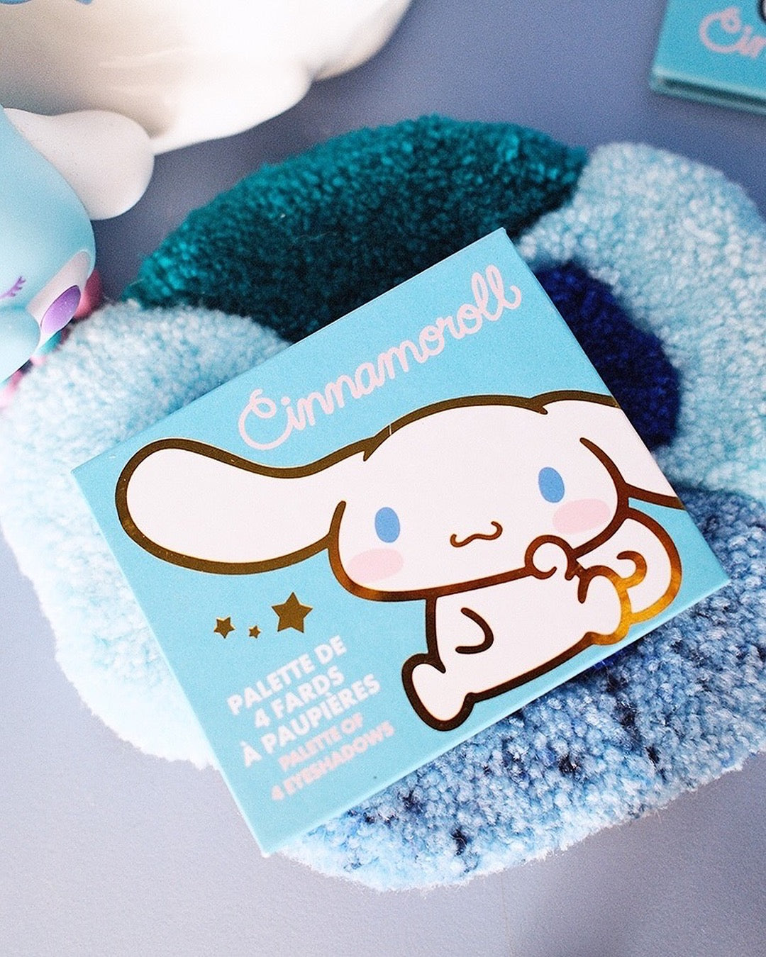 Une palette de fards à paupières Sanrio Cinnamoroll à 4 couleurs ouverte, comprenant des teintes mates et irisées de bleu, gris foncé, brun et argent, avec un applicateur double embout et un miroir intégré.