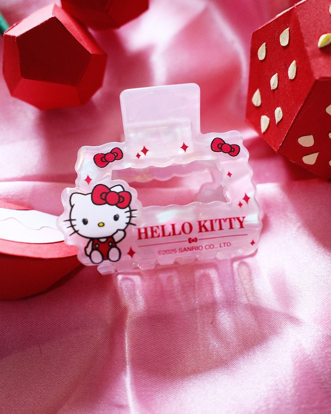 Petite pince à cheveux nacrée Hello Kitty posée à plat sur un tissu satiné rose à côté de fraises décoratives en papier.