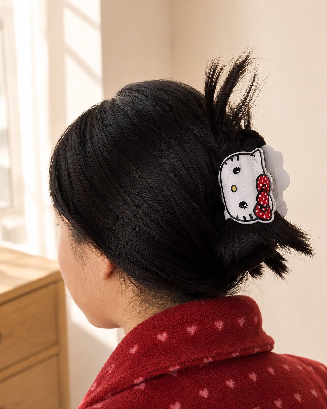 Vue arrière des cheveux bruns d’une femme attachés en chignon à l’aide de la pince à cheveux blanche en forme de visage Hello Kitty, portée avec un peignoir rouge.
