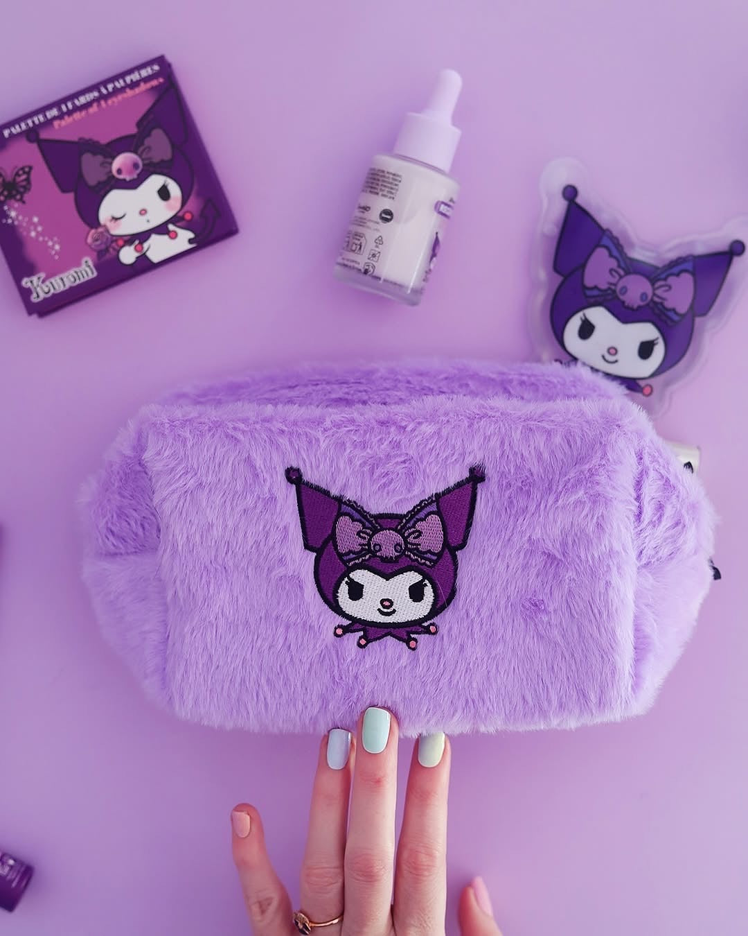 Une main présentant une trousse de maquillage en peluche fausse fourrure violette de  Kuromi Sanrio avec d'autres produits cosmétiques de la même collection.