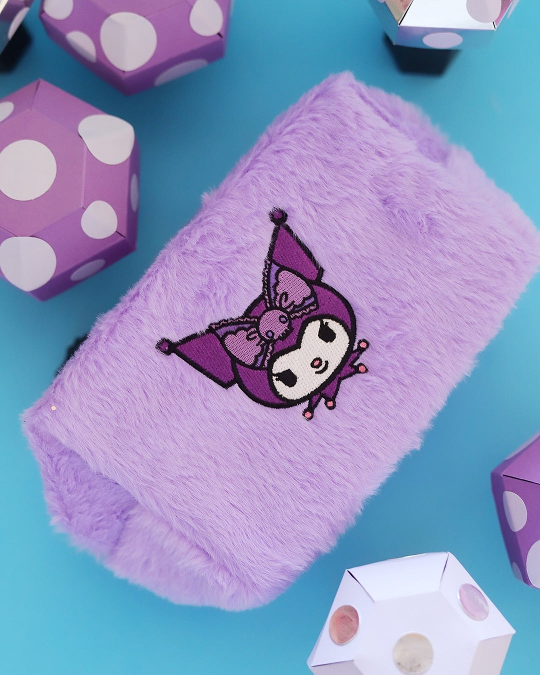 Vue de biais et de dessus d'une trousse cosmétique en peluche fausse fourrure violette Kuromi entourée de décors géométriques à pois violet et blanc sur fond turquoise.